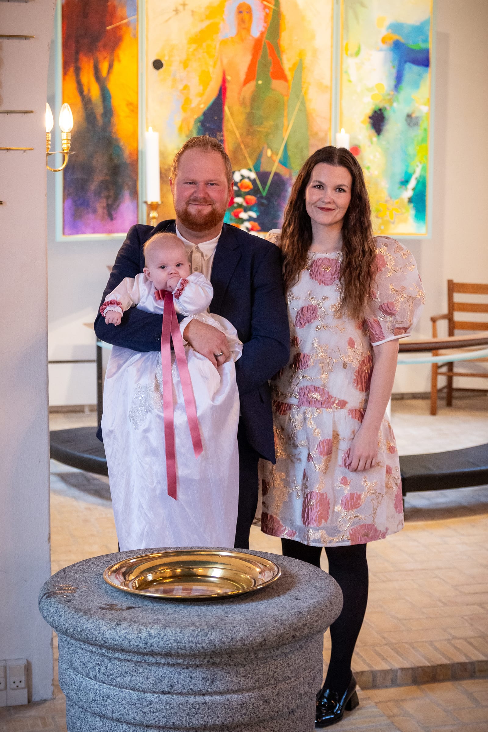 Forældre holder deres baby i dåbskjole ved døbefonten i kirke