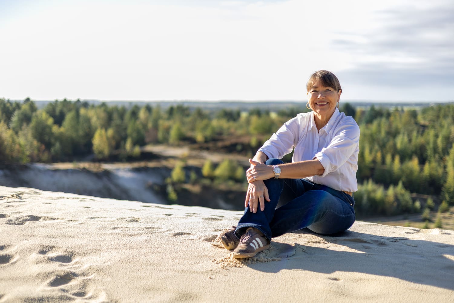 Smilende kvinde i hvid skjorte sidder i sandet på en klit med udsigt over skov